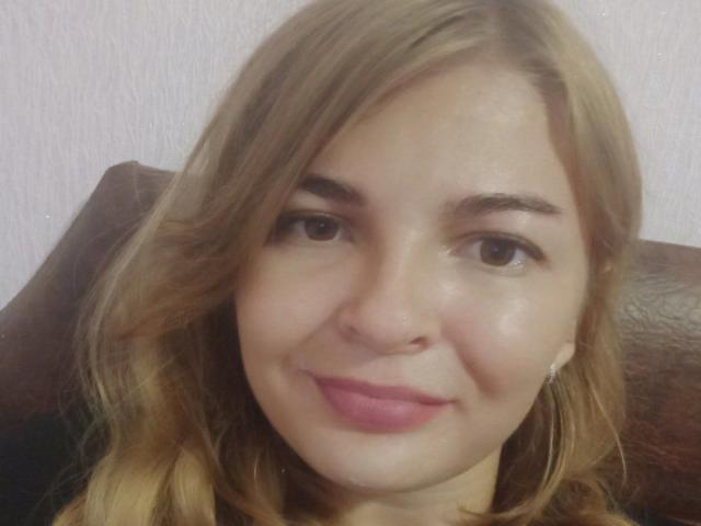 RoxannaSonia has light brown hair, brown eyes, pink lipstick, and a slight smile. She's wearing small earrings and sitting against a brown leather chair.