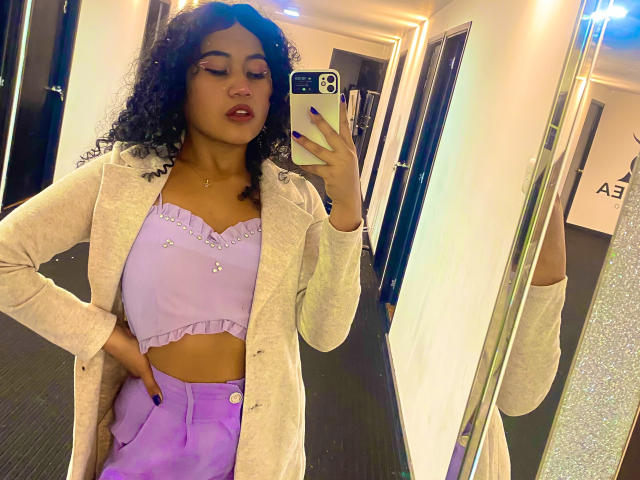SophieSonyas with curly black hair, purple crop top, and buttoned purple pants, wearing a beige coat, taking a mirror selfie in a well-lit, modern room.