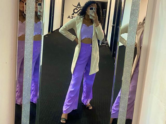 SophieSonyas, gekleed in een paarse broek en top, met een beige vest, staat voor een spiegel en houdt een telefoon vast. In de spiegel is haar reflectie te zien, op een zwarte vloerkleed, met bruine sandalen en krullend blauw haar..