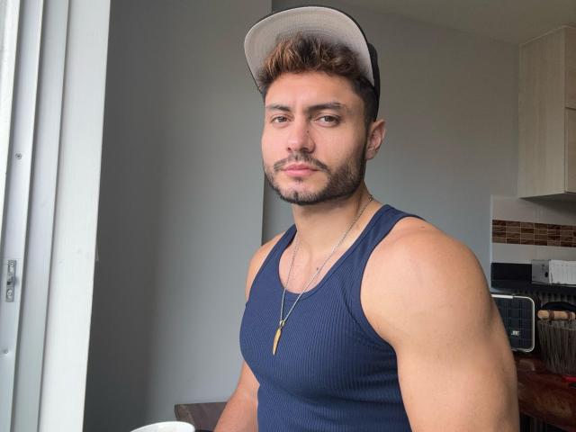 ManuClark with a muscular build, wearing a navy tank top, beige cap, and necklace with a gold pendant, looking at the camera with a slight smirk.