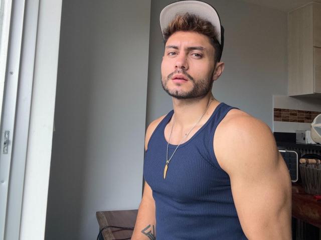 ManuClark with brown hair, beard, and cap, wearing a blue ribbed tank top, showing muscular arms and a tattoo, standing by a window, looking at the camera.
