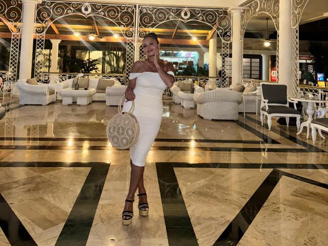 GalaMalkova stands in a marble-floored room, wearing a tight white off-shoulder dress, black heels, and a round wicker purse, with her hand touching her chin.