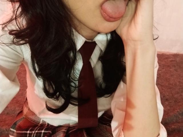 LexyFoxy69, avec de longs cheveux noirs, la langue tirée, touchant sa joue, portant une chemise blanche, une cravate rouge et une jupe à carreaux, sa main près de son visage..