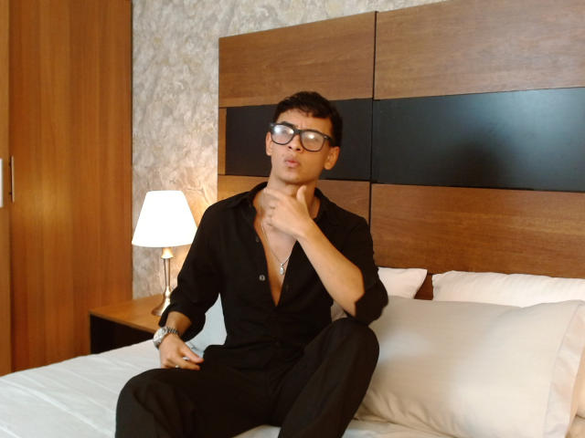 Brian O'Connor, con gafas, cabello oscuro y corto, camisa negra, pantalones negros, sentado en la cama, tocándose el cuello, almohada blanca, cabecera de madera, panel negro, lámpara a la izquierda, pared blanca..