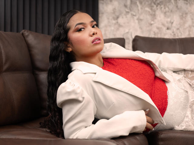 NathaliaBrownn is laying on a brown leather couch, wearing a white jacket over a red lace top, looking up with a slightly open mouth, long black wavy hair.