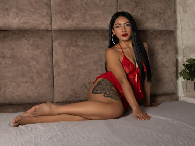 LillyTaylor with long black hair, red lace lingerie, and a tattoo on her right thigh, sitting on a bed, legs crossed, looking at the camera with red lipstick.