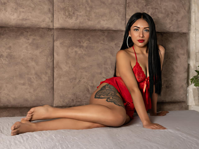 LillyTaylor, with long black hair, red lace lingerie, and a tattoo on her right thigh, sits on a bed, legs crossed, showing her bare ass and small tits.
