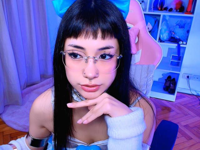SabriPrettty has long black hair with blue bow, glasses, nose ring, and white sleeve, touching her chin, showing off her small tits and cute pink lips, in a blue-lit room.