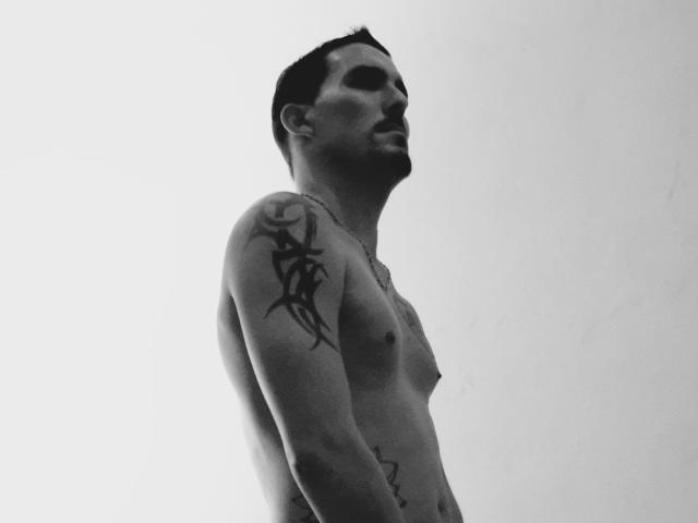 ExedeNieve, shirtless, standing, facing right, showing a black tribal tattoo on his right shoulder, short dark hair, slight mustache, grayscale, high-contrast, slight shadow on his chest.