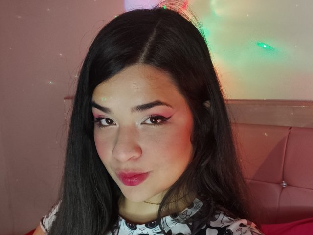 AntonelaPark with long black hair, pink eyeliner, red lipstick, and a subtle smile, looking at the camera with a cute, sexy face, wearing a patterned top.