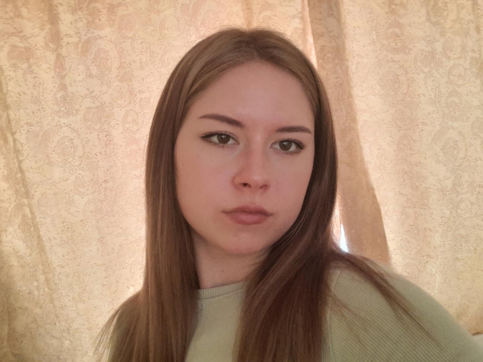 TomyRisyuki has long brown hair, light skin, and is wearing a green ribbed shirt, looking straight at the camera with slightly pouty lips and thin black eyeliner.
