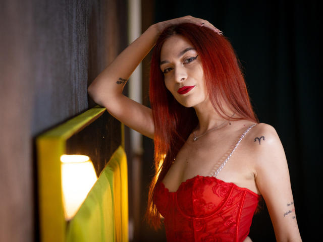 KloeyAdais con cabello rojo, corpiño de encaje rojo, tirantes de perlas, brazo levantado, mano en la cabeza, tatuajes, lápiz labial rojo, mirando a la cámara, sonrisa sutil, iluminación suave, fondo oscuro..
