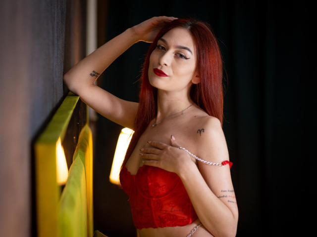KloeyAdais met rood haar, een rode kantbeha en rode lippenstift, met één hand op haar hoofd en de andere hand op haar borst, tatoeages op haar arm, staand naast een geelgroene muur, donkere achtergrond..