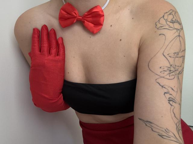 EricaLust con guanti rossi, top nero senza spalline, fiocco rosso e braccio tatuato. Il suo seno è visibile, mostrando un po' di scollatura. La sua mano è vicino al petto..