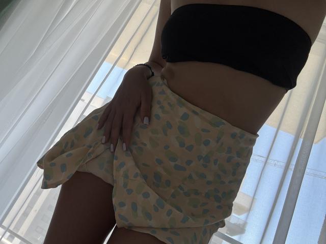 EricaLust with a black strap top, wearing a yellow skirt with blue polka dots, exposing white panties, standing in front of sheer curtains, showing her flat stomach and hand.
