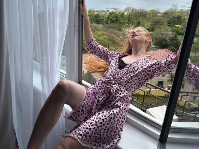 GingerFairy, met lang rood haar, gekleed in een roze jurk met luipaardprint, leunt tegen een raam, met armen omhoog en benen gebogen, kijkt naar buiten, met een lichte glimlach, terwijl de wind door haar haar waait..