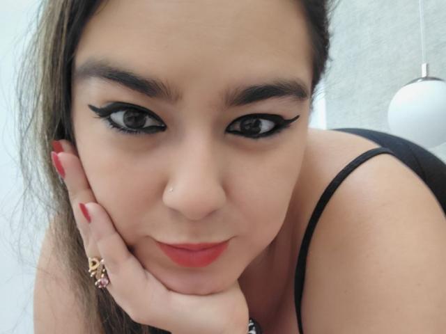 TaylorNova con delineador negro, lápiz labial rojo y uñas rojas, inclinada hacia adelante, con la mano en la mejilla, vistiendo una blusa negra, un piercing en la nariz y un anillo con piedras rosas..