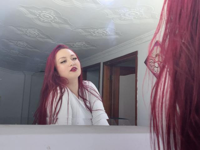 FreyaRose with red hair, white top, looking in mirror, reflection shows her face and red hair, subtle nose piercing, slightly open mouth, white ceiling with ornate patterns.