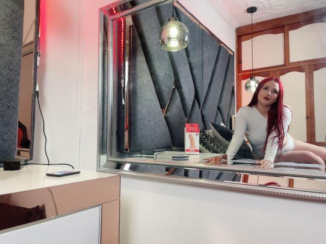 FreyaRose with red hair, white long-sleeve top, and black shorts, leaning on a mirror, reflecting her tits, ass, and face, with a metallic hanging light above.