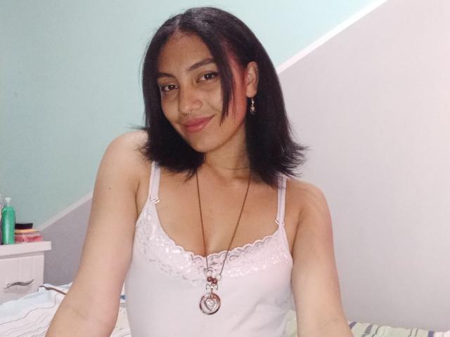 KanyaLopez with shoulder-length black hair, smiling, wearing a white lace-trimmed tank top, necklace, and earrings, showing her tits. Bedroom background with green and white walls.