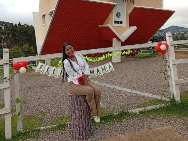 MariamLara, com longos cabelos pretos, sorrindo, vestindo uma blusa branca, calças marrons e uma saia estampada, sentada em um banco de madeira branca, segurando um pompom vermelho..