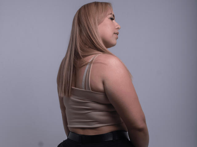 ZaharaBennet has blonde hair, a beige tank top, and black pants, showing her back and side profile with a slight smile, her butt visible in the shot.