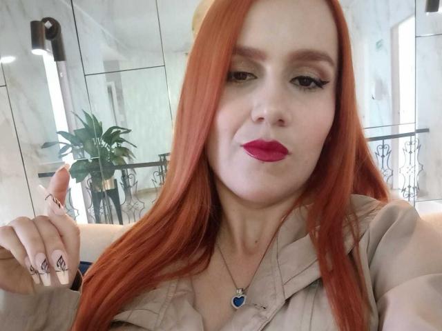 SelenneMillers with red hair, wearing pink lipstick, giving a thumbs-up, showing a heart necklace, and a tattoo on her ring finger. Boobs visible under a beige shirt.