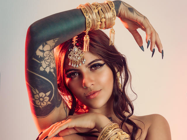CoriPaige with dark tattoos on arm, wearing gold jewelry, including bangles and a headpiece, has long nails and brown wavy hair, looking at camera with a slight smile.