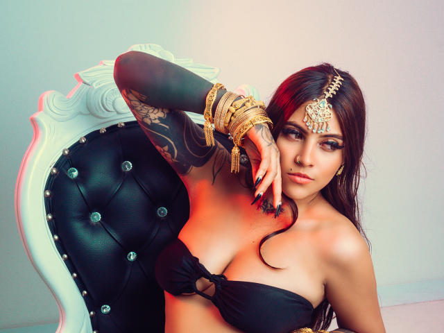 CoriPaige con i capelli scuri, una top nera senza spalline che mostra il seno, gioielli dorati, un braccio tatuato, unghie nere, seduta su una sedia bianca con rivestimento nero, che guarda la fotocamera..