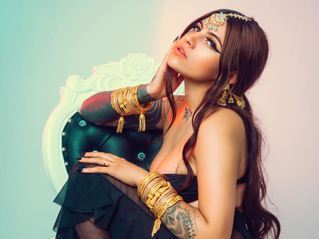 CoriPaige with long brown hair, wearing gold jewelry and a black dress, sits on a green velvet chair, hand on chin, tattoos visible, looking up with a sultry expression.