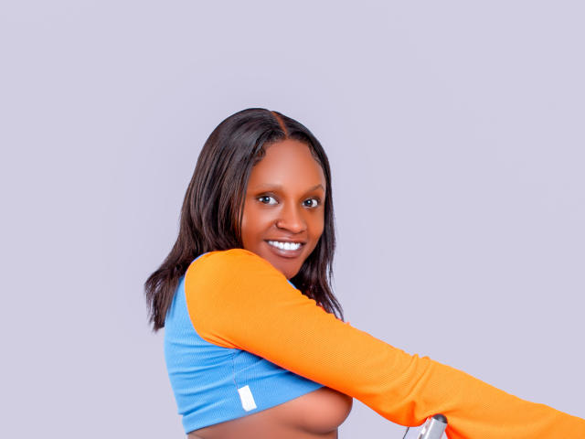 SexyMhyrah with dark skin, smiling, wearing a blue and orange long-sleeve top, exposing her tits, brown hair, and white teeth, against a plain background.