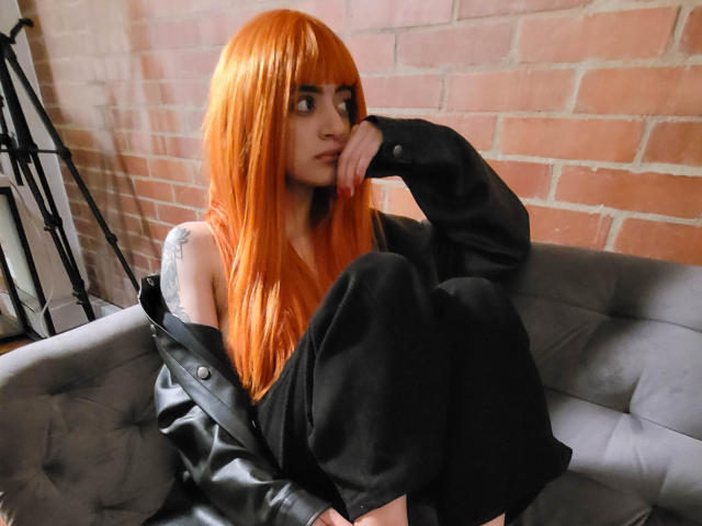 Amelie Sato mit orangefarbenen Haaren, schwarzem, schulterfreiem Oberteil, schwarzen Hosen, Tattoo am rechten Arm, sitzt auf einem grauen Sofa, die Hand am Kinn, und blickt zur Seite. Im Hintergrund ist eine Ziegelwand zu sehen..