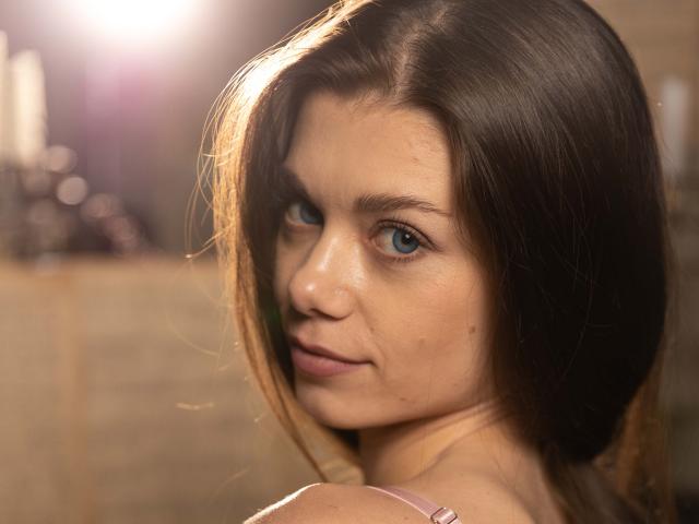 EvaMcCoy has blue eyes, brown hair, and a slight smile, looking over her shoulder with soft light highlighting her face. She has small tits and a cute nose.