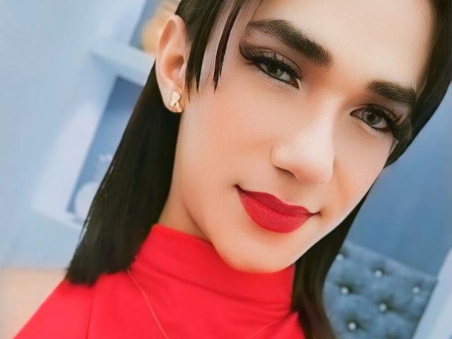 KatrinaQueeny with red lipstick, dark eyeliner, and a red turtleneck, has straight black hair, small hoop earrings, and a cute smile, looking directly at the camera.