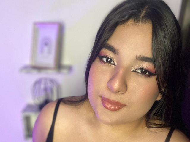 AuroraSxx with shiny brown eyes, dark straight hair, and glossy red lips, wearing a black spaghetti strap top, looking slightly to the side with a subtle smile.