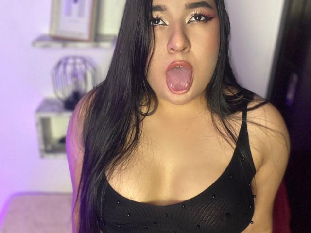 AuroraSxx with long black hair, eyes lined with pink and black makeup, tongue out, wearing a black, sparkly tank top, exposing her boobs.