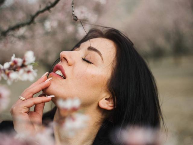 Annilay's face, eyes closed, dark hair, pink lips, hand near mouth with long nails, cherry blossoms in focus, soft pink and white, gentle expression, ring on finger.