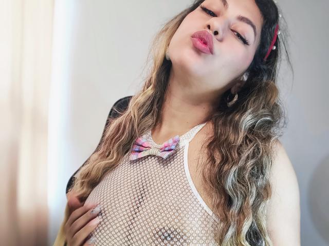 AnastaciaMonaco with puffy pink lips, wavy blonde hair, wearing a white mesh top, pink bowtie, and black sleeves, holding her tits, looking down with half-closed eyes.