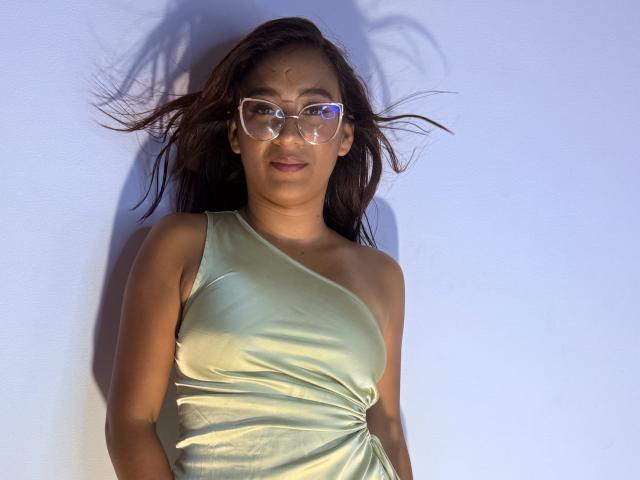AnjaliKhushi with light green satin dress, clear glasses, and brown hair blowing, standing against white wall, showing small tits and cute face.