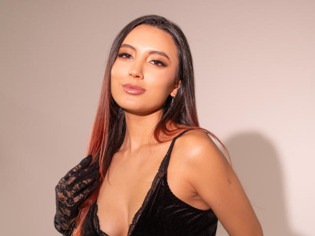 CarlaFranco has long brown hair with red tips, wearing a black lace dress with a deep cleavage, showing her shiny, pinkish lips and a confident, sexy expression.
