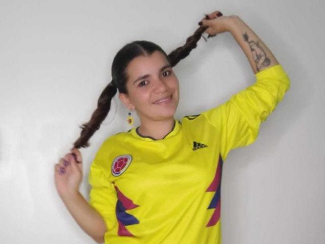 BulmaHot with braided hair, holding her hair up, smiling, wearing a yellow soccer jersey, purple and red stripes, visible tattoo on her right arm, white background.
