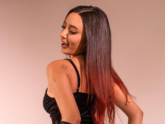 CarlaFranco con lunghi capelli lisci, di colore nero sfumato in rosso, indossa una canottiera nera, sorride, mostrando piccoli tatuaggi sulla spalla, e guarda oltre la spalla, con uno sfondo rosato..