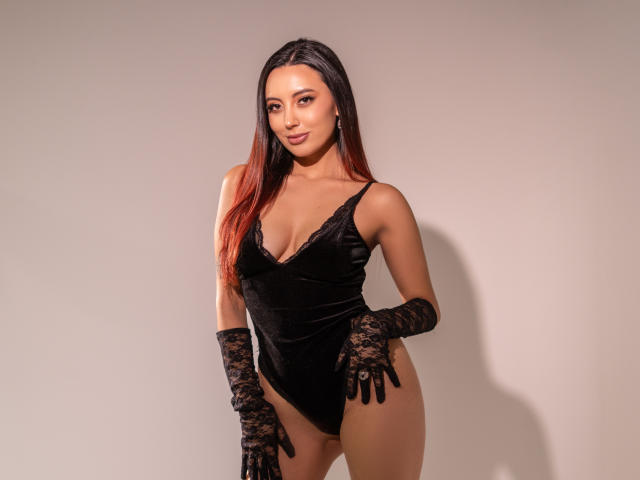 Carla Franco, con cabello largo de color rojo y negro, guantes de encaje negro y un body de terciopelo negro que muestra sus pechos y vulva, está de pie frente a un fondo liso, sonriendo ligeramente..