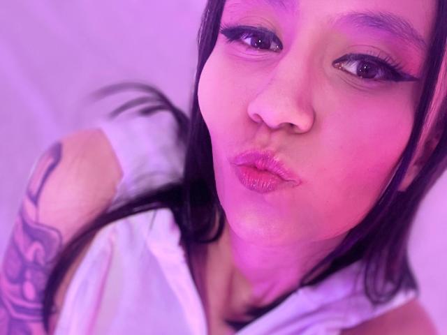 EviieGothel with pink-tinted skin, pouty lips, and long eyelashes, wearing a white shirt, showing a tattoo on her right arm, looking at the camera.