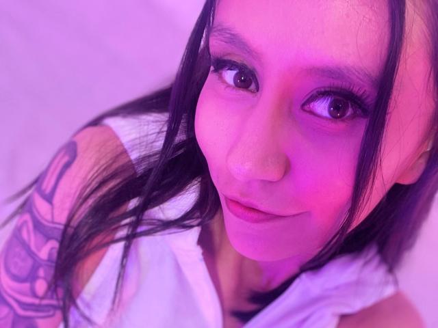 EviieGothel's face with purple lighting, long black hair, tattoo on right arm, brown eyes, slight smile, white shirt, nose ring, and pinkish-purple skin tone.