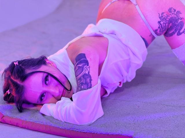 EviieGothel is on her knees with her ass up, wearing a white shirt and pink thong, showing tattoos on her arm and thigh, with purple lighting.