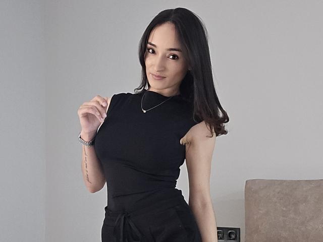 CandyLoves with black dress, sleeveless, medium tits, dark brown hair, subtle smile, necklace, bracelet, tattoo on right arm, standing, gray wall, beige chair in background.