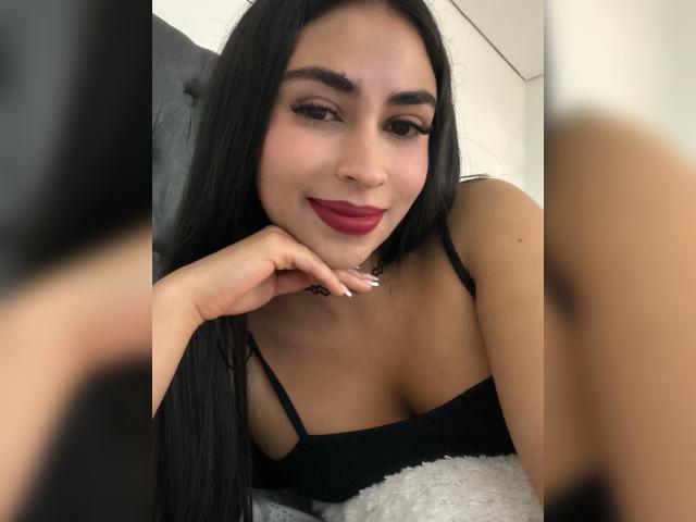 MiilaWx with black hair, red lipstick, and black strap top, resting her chin on her hand, showing her cleavage and a cute smile, in a mirror selfie.