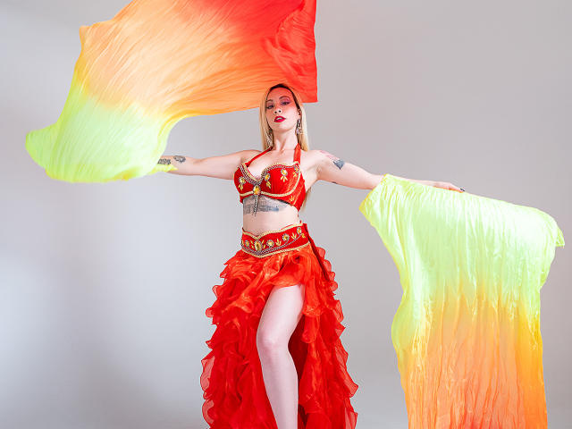KaatArt with orange-red and yellow-green flowing fabric, wearing a red bra with gold star and red ruffled skirt, showing her tits and ass, tattoos on arms, white background.