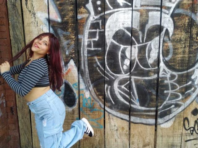 SophieHill with red hair, striped top, and high-waisted jeans, standing against a graffiti-covered wooden wall, kicking one leg up, showing her ass.